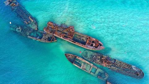 Aerial view of several shipwrecks resting in shallow, turquoise waters, their rusted remains a stark contrast to the vibrant sea.