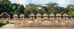 A row of traditional huts with thatched roofs sits atop a rocky outcrop, backed by lush green trees under a partly cloudy sky.