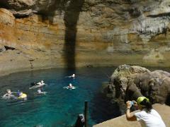 People swim in the crystal-clear water of a cavern while one person on a rock outcropping takes a photo.