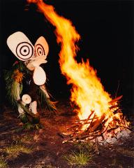 A person in a large, eye-catching mask and costume stands near a large bonfire, likely as part of the Rabaul Mask Festival and Baining Fire Dance in Papua New Guinea.