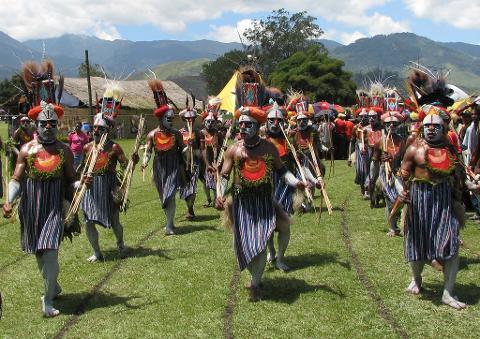 A group of men in traditional tribal clothing and face paint parade on a grassy field during the Mt Hagen Show.