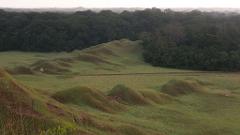 Rolling grassy hills are dotted with small mounds in a vast landscape with a dense forest in the background.