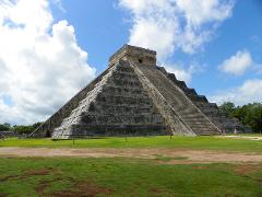Chichen Itza Airplane Tour and Day Visit from Playa Del Carmen An upward shot captures the majestic Chichen Itza pyramid, towering over lush green grass under a partly cloudy blue sky.