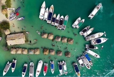 Una vista aerea mostra un&#39;isola tropicale molto frequentata, con un gruppo di capanne con il tetto di paglia immerse in acque turchesi poco profonde, circondate da numerosi yacht e barche, brulicanti di persone che si godono le acque assolate.