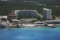 This is an aerial view of a resort in Cozumel, Mexico, showcasing a large building with multiple floors, a pyramid-shaped thatched-roof structure, a white hotel building, a beach with umbrellas, and clear blue water with a boat.