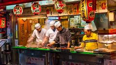Four cooks in traditional hats prepare food at a bustling Osaka street food stall, illuminated by vibrant lanterns with Japanese characters.