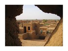 Certainly! Here's a concise description of the image:A view of a tightly packed earthen-built village or town is seen through an irregular earthen frame or window, showcasing the architecture characteristic of a desert region.