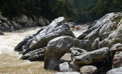 Kayak Adventure - 10 Days A kayaker braves the rapid current of a river, surrounded by large, gray rocks, with lush green foliage in the background.