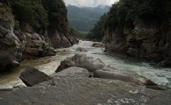 Kayak Adventure - 10 Days A kayaker navigates a river flanked by rocky cliffs and lush vegetation in a valley surrounded by mountains under a cloudy sky.