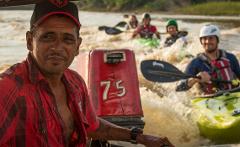 Kayak Adventure - 10 Days A man in a red plaid shirt and baseball cap is piloting a boat, with kayakers paddling in the turbulent water behind him.