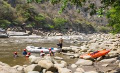 A group of people in helmets and life jackets are in a river near a raft and a kayak, with a man standing on the raft and a rocky, wooded hillside in the background.