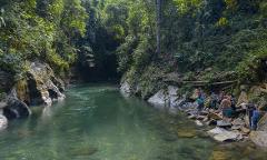 A scenic river winds through lush greenery as people gather on the rocky bank, enjoying the natural beauty.