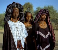 Three Wodaabe women stand outdoors in traditional attire, including colorful headwraps, necklaces, and facial markings, against a backdrop of desert vegetation and a clear sky.