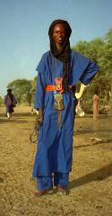 A Tuareg man in a blue robe, head scarf, and belt with decorative items stands in a dry, open landscape with trees, camels, and other figures in the background.