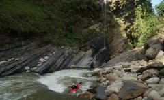 Kayak Adventure - 10 Days A kayaker in a red kayak navigates a turbulent river with a waterfall and rocky cliffs in the background.