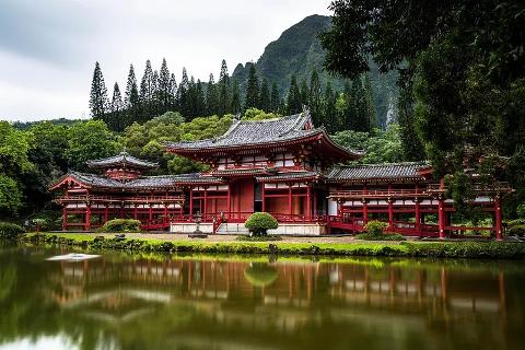 The red Byodo-In Temple is reflected in the calm water of a pond surrounded by lush greenery, with a backdrop of forested hills and a cloudy sky.