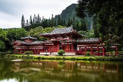 The red Byodo-In Temple is reflected in the calm water of a pond surrounded by lush greenery, with a backdrop of forested hills and a cloudy sky.