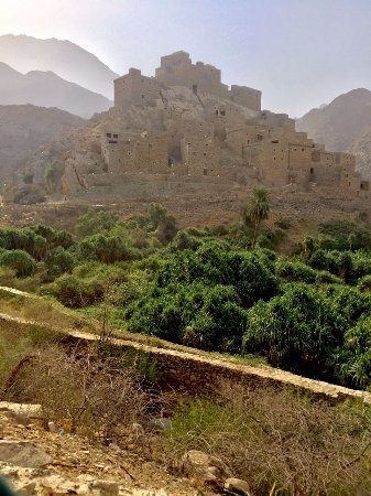 A medieval-style fortress town sits atop a rocky hill, surrounded by green vegetation and mountains in the background, creating a landscape of historic charm and natural beauty.