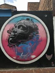 A colorful mural on a brick wall depicts a person's face in a circular frame, blending black, pink, and blue hues.