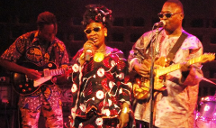 Three African musicians are on stage, a woman in a patterned dress sings into a microphone, while two men on either side of her play electric guitars.