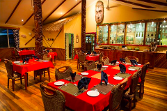 This is a warm-toned interior shot of the Tawali resort dining room, featuring wooden floors, tiki-style carvings, tables set with red cloths, woven chairs, and a well-stocked display cabinet.