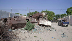 The image shows a derelict armored vehicle wreckage on a dusty street, likely a remnant of conflict, with a pickup truck carrying armed individuals passing by.
