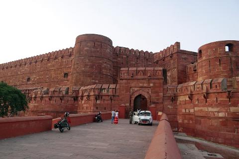 A view of Agra Fort's imposing red sandstone walls, battlements, and gate, with people and vehicles in front.