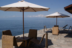 Ein malerischer Blick auf eine Terrasse am Wasser mit geflochtenen Stühlen und Tischen unter großen Sonnenschirmen und Blick auf ein ruhiges Gewässer mit Hügeln in der Ferne.