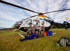 A group of people poses in front of a white and blue helicopter with its rotors, parked on a grassy field under a partly cloudy sky.
