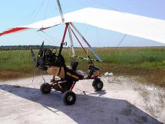 The image shows an ultralight aircraft parked on a sandy area, with a field of tall grass in the background under a bright, sunny sky.