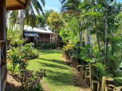 A vibrant garden path leads to a cozy wooden house, surrounded by lush tropical greenery and palm trees, creating a tranquil and inviting scene.
