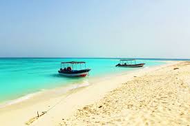 Two boats are anchored near a white sandy beach with crystal clear turquoise water under a clear blue sky.