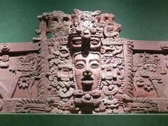 This image is a detailed reddish-brown stone carving of a Mayan deity or ruler, adorned with intricate symbols and surrounded by decorative elements, against a dark green background.