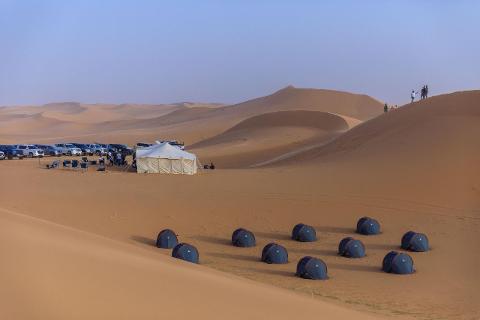A desert landscape with a campsite, tents, cars, and people on a dune.