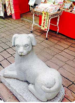 En stenstatue af en hund står på et offentligt sted, sandsynligvis i nærheden af en butik eller et marked, med varer udstillet i baggrunden.