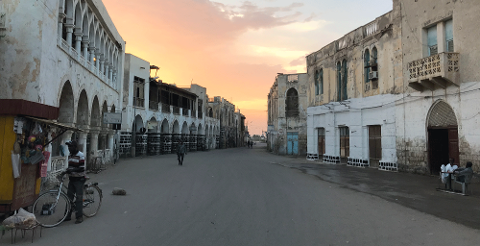 An old street at sunset with dilapidated buildings, a merchant with a bicycle, and men sitting by a doorway.