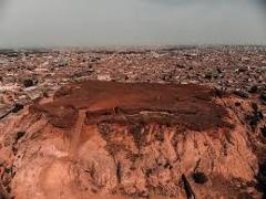 Here's a one-sentence description of the image:This aerial shot depicts a reddish-brown plateau-like formation with eroded sides, situated in the middle of a densely populated urban area in Kano, Nigeria.