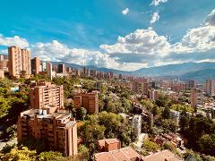 A high-angle cityscape with red brick buildings nestled among lush greenery under a bright blue sky with puffy white clouds, and a mountain range in the distance.