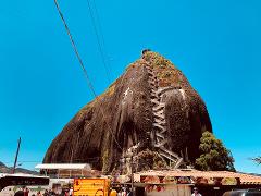 A giant rock with a long staircase winding up its side stands tall against a clear blue sky, surrounded by small shops and buses, depicting a bustling tourist attraction.