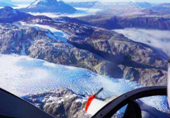 An aerial view shows a vast glacier snaking through rocky, mountainous terrain, partially obscured by mist and framed by the cockpit of an aircraft.