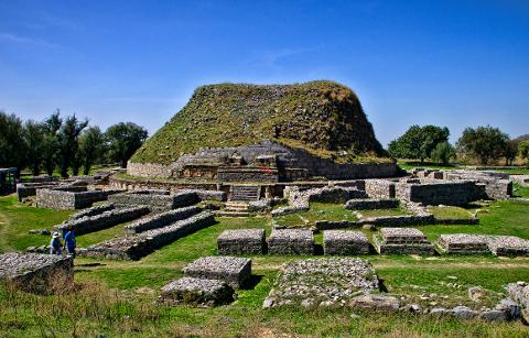 An ancient stone stupa in Taxila, Pakistan, surrounded by green grass and ruins of what appears to be walls, set against a clear blue sky.