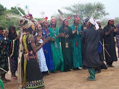 A group of people in traditional clothing, including elaborate headdresses, are gathered outdoors, with some clapping and one appearing to dance.