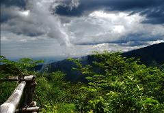 A lush green mountain vista is framed by a bamboo railing, while a striking cloud formation descends from a dramatic, stormy sky.