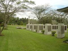 A serene view of Rabaul War Cemetery, with headstones standing in neat rows on a green lawn, under trees, a war memorial in the background.