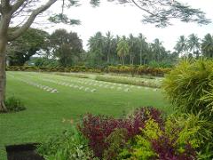 A serene cemetery with rows of headstones on a lush green lawn, bordered by vibrant flowers and tropical trees, creates a peaceful memorial scene.