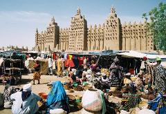 A bustling market scene unfolds in front of the Great Mosque of Djenné, with vendors and shoppers engaging in commerce and everyday life.