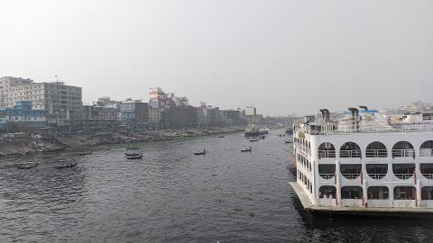 3-Day Dhaka Heritage & Culture Tour — Old Palaces, Lost Cities & Zamindari Mansions A wide river flows past a bustling cityscape of buildings and a large white ferryboat, with smaller boats dotting the water.