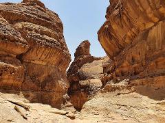 A narrow canyon passage carved through textured sandstone cliffs under a bright sky.