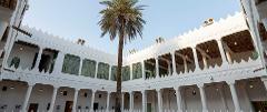 A low-angle shot reveals a white building with sawtooth-edged roofs and columns surrounding a courtyard with a tall palm tree in the center.