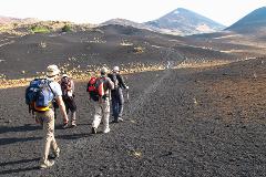 A group of hikers walks across a black, rocky landscape towards distant mountains under a clear sky.